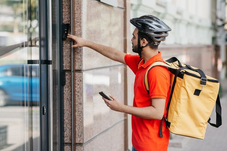 Un homme portant un casque et une chemise orange tient un sac, debout avec confiance dans un décor extérieur.